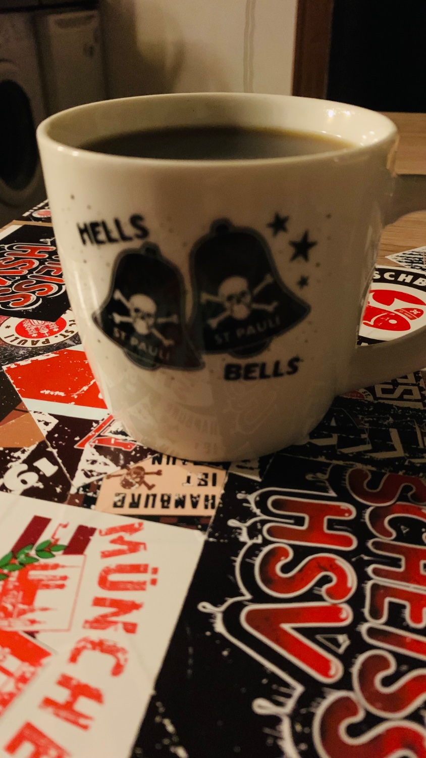 Eine Kaffeetasse mit dem Aufdruck „HELLS BELLS“ und Totenkopfmotiven ist mit dunklem Kaffee gefüllt. Die Tasse steht auf einem Tisch, der mit verschiedenen bunten Aufklebern und Grafiken bedeckt ist.