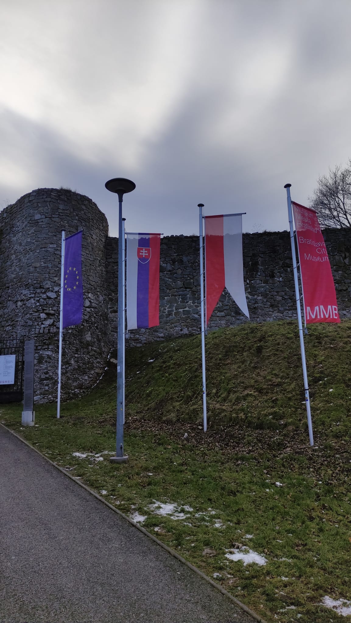Four flags hoisted vertically, each on their own flagpoles. From left to right, those flags are of: European Union, Slovakia, Bratislava and Bratislava City Museum. In front of the image, a part of the asphalt can be visible. Behind it is a grass surface followed by a gray stone wall behind the flags.