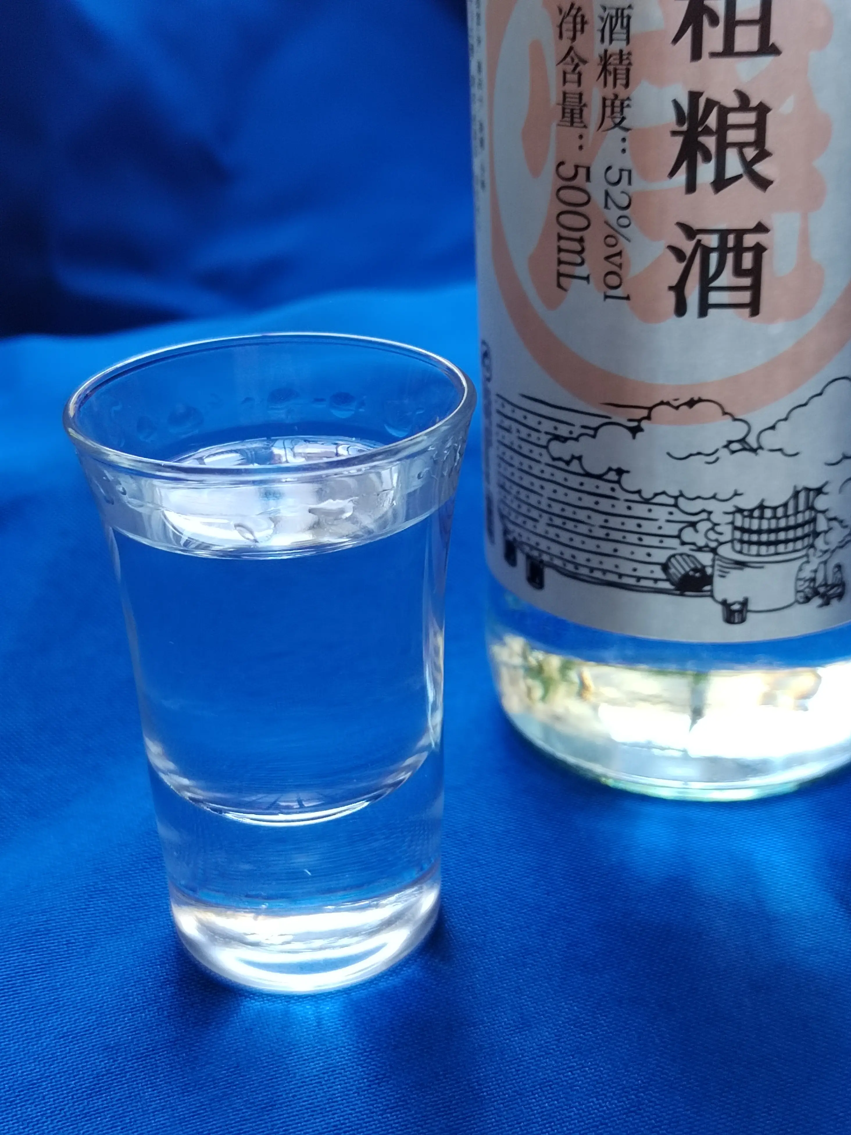 A shot glass with a transparent liquor displaying ethanol tears in abundance in the foreground with the bottle it came from behind it in the background.  The bottle's only non-Chinese text says "500ml" and "50%vol".
