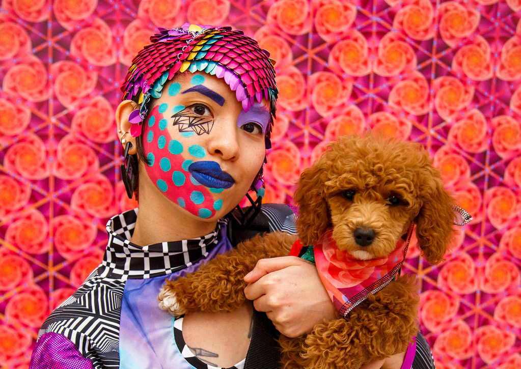 Person from the chest up, holding a small curly haired brown dog. They are wearing a metallic multicolored scaled flat headdress that comes to a point at their forehead. Their face is painted with a swooping line of orange on their jawline, dotted with circles of light blue, and their dark blue lipstick is asymmetric, with one half of their upper lip overdrawn. Their shirt is a pachwork of multiple patterns of check, galaxy print, and pink snakeskin print, with a mock turtleneck and a circle cutout in the upper chest.