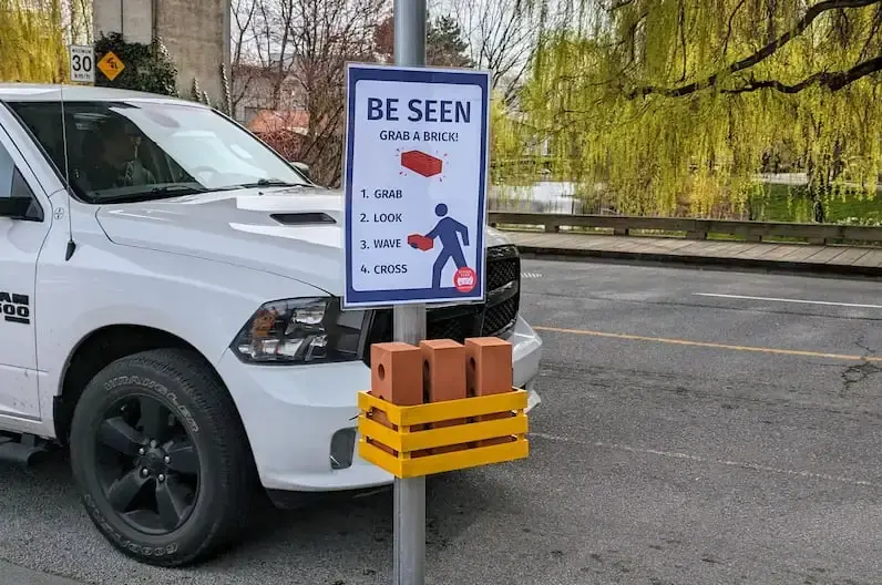 yellow box containing three bricks with a sign that says "BE SEEN Grab a Brick 1. Grab 2. Look 3. Wave 4. Cross"
