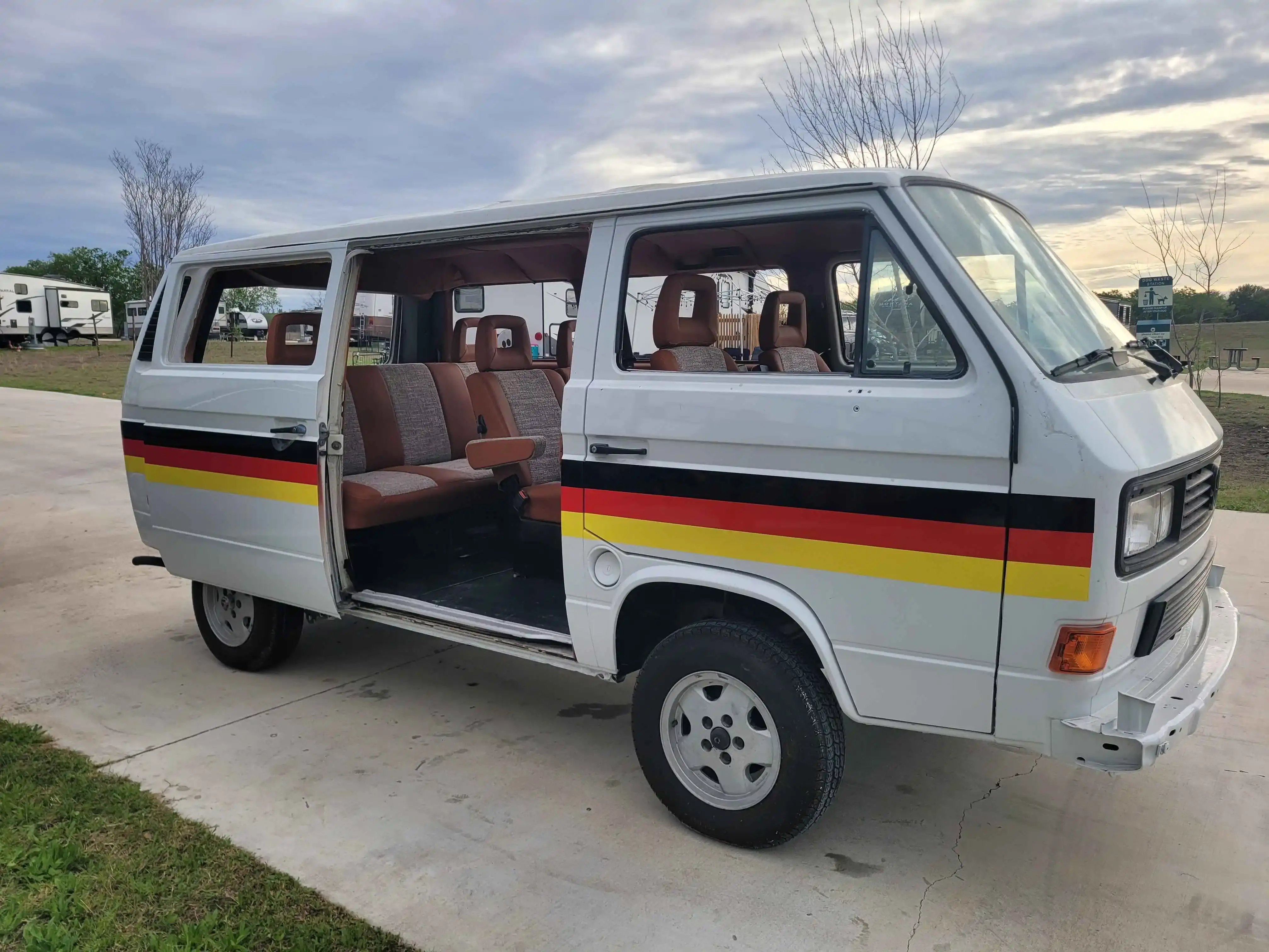 A 1990 Volkswagen Vanagon painted white with stripes in the colors of the German flag running down the sides