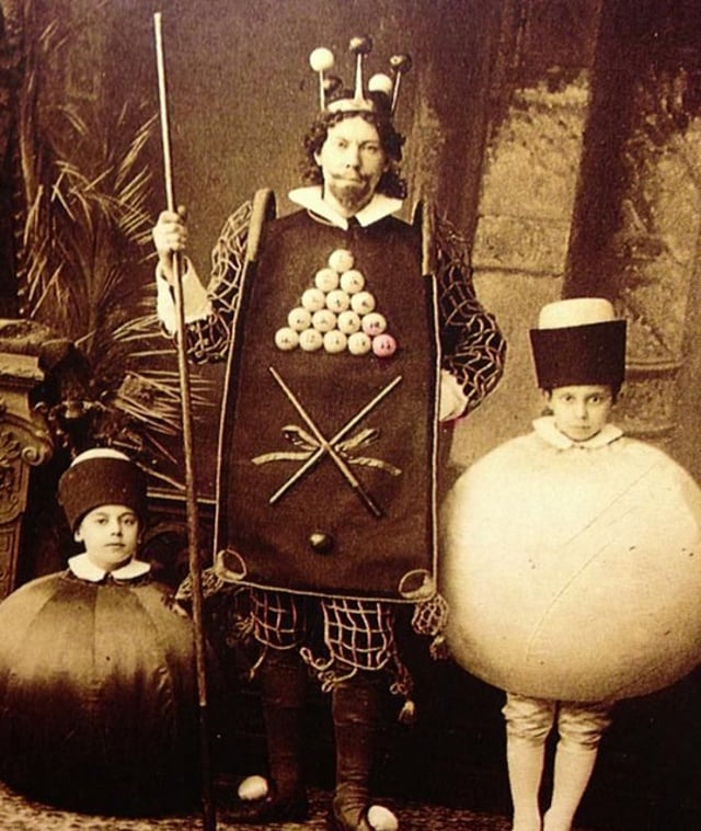 A black and white photo of a man with a tabbard featuring pool balls and cues, holding a pool cue, and flanked by two children dressed as pool balls. 