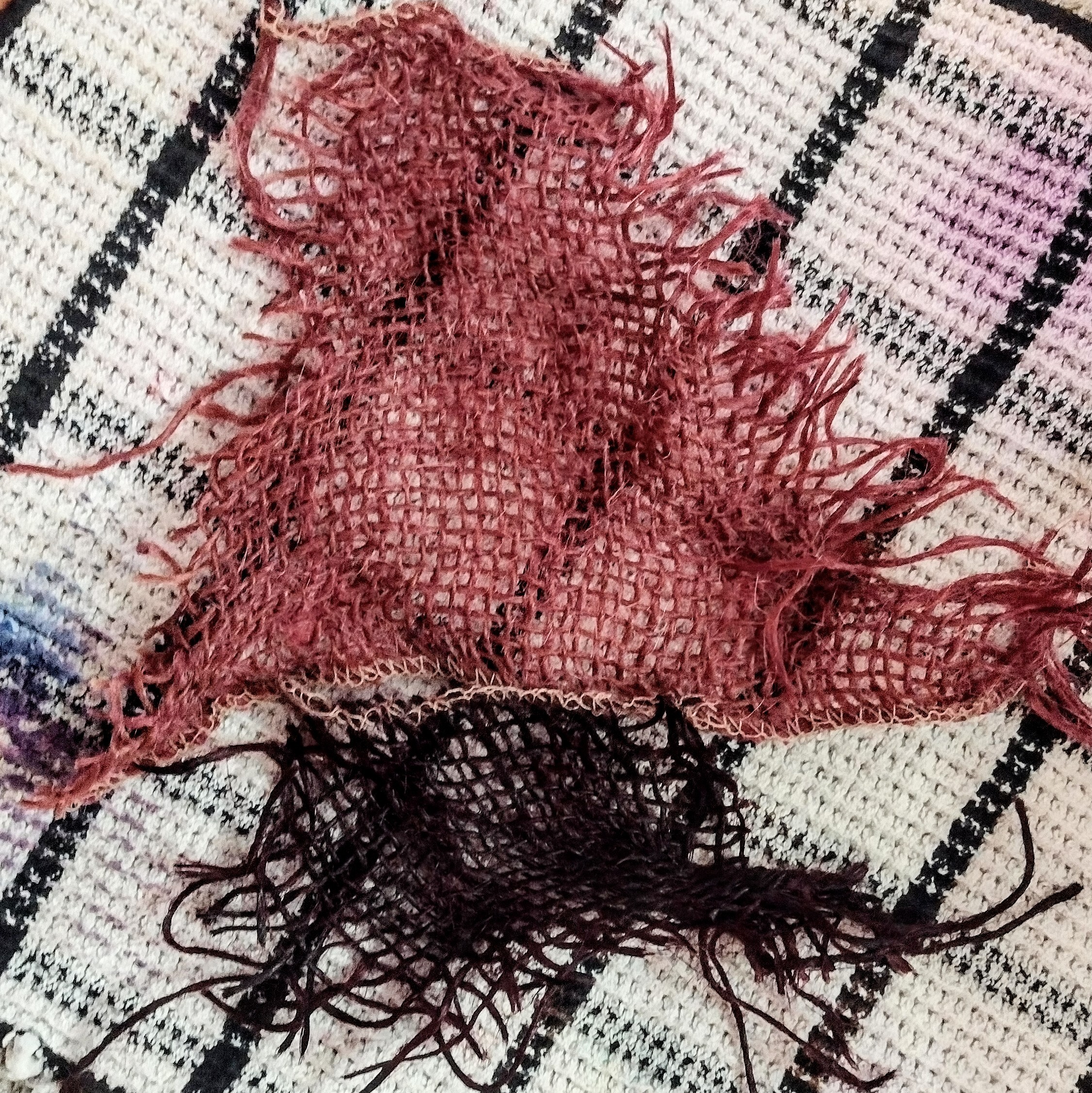 Two scraps of loose woven burlap on a stained dishcloth. The upper scrap is a light wine red and the lower, smaller scrap is a deep, almost black red.