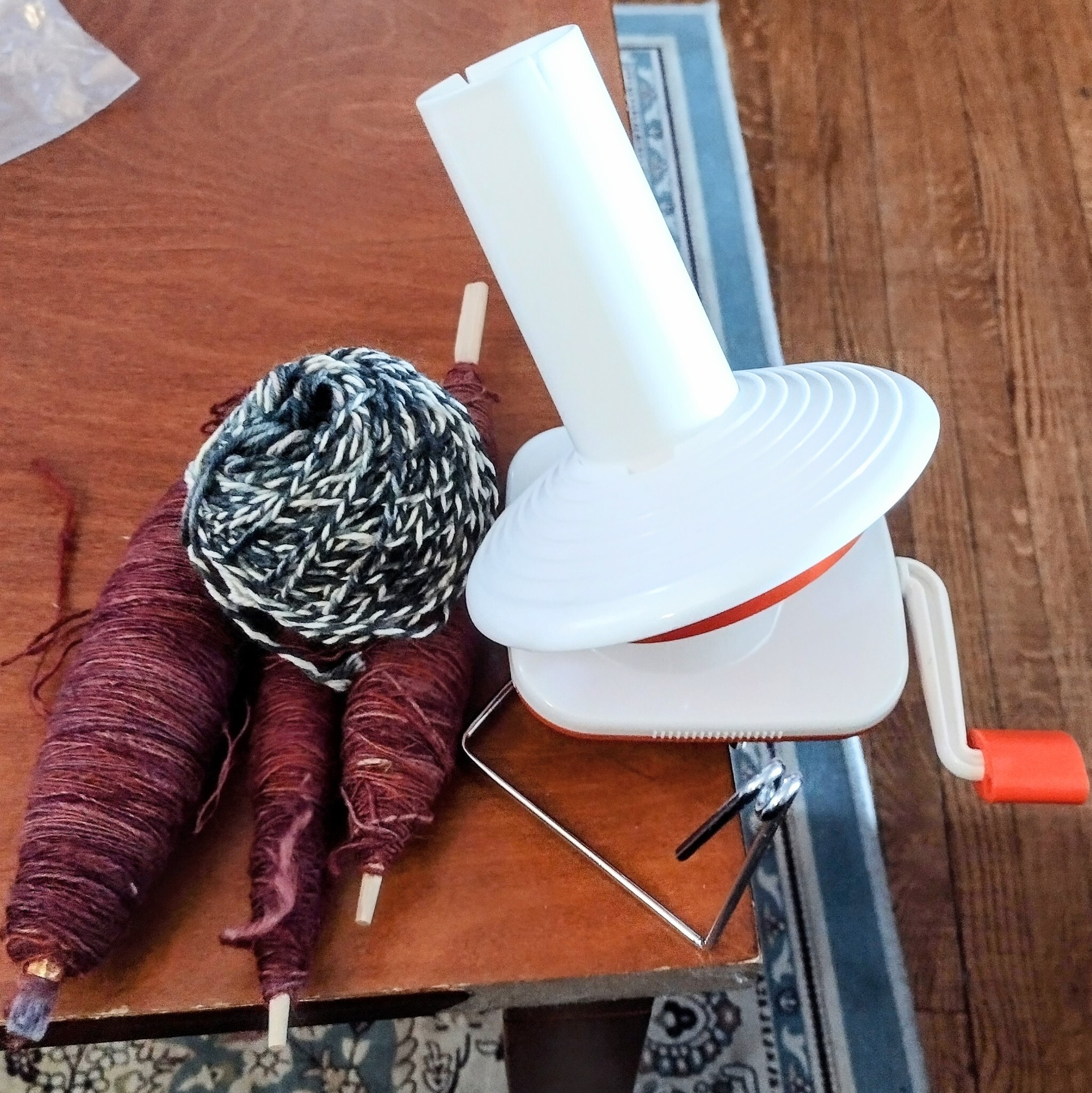 A manual yarn ball winder attached to the side of a table, next to a freshly spun ball of gray and white yarn, sitting on three spindles of red thread.