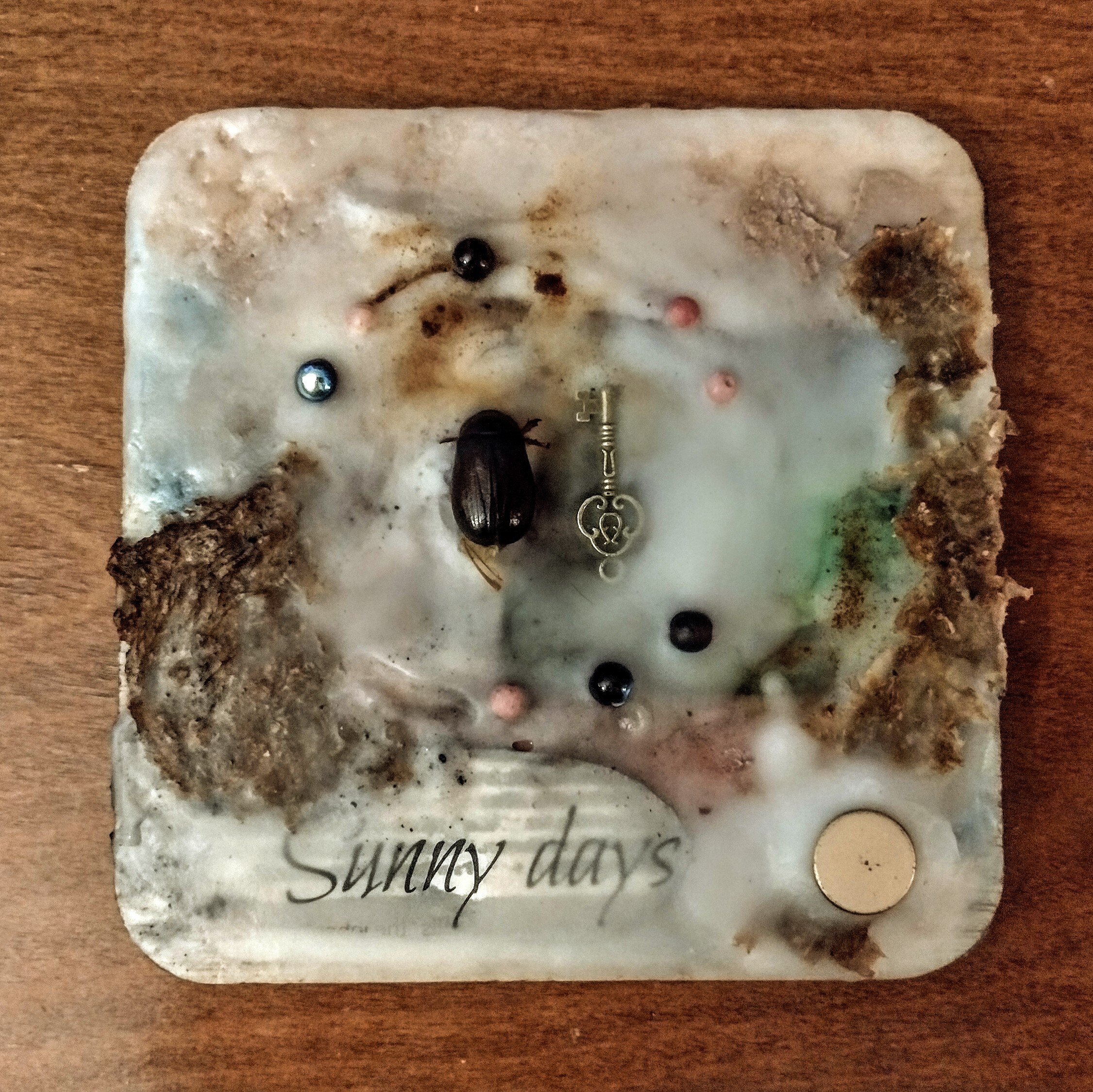 A four-inch-square mixed media artwork including cut up magazines, handmade paper, a dead june bug, beads and a small key all set in encaustic wax medium with powdered pigments.