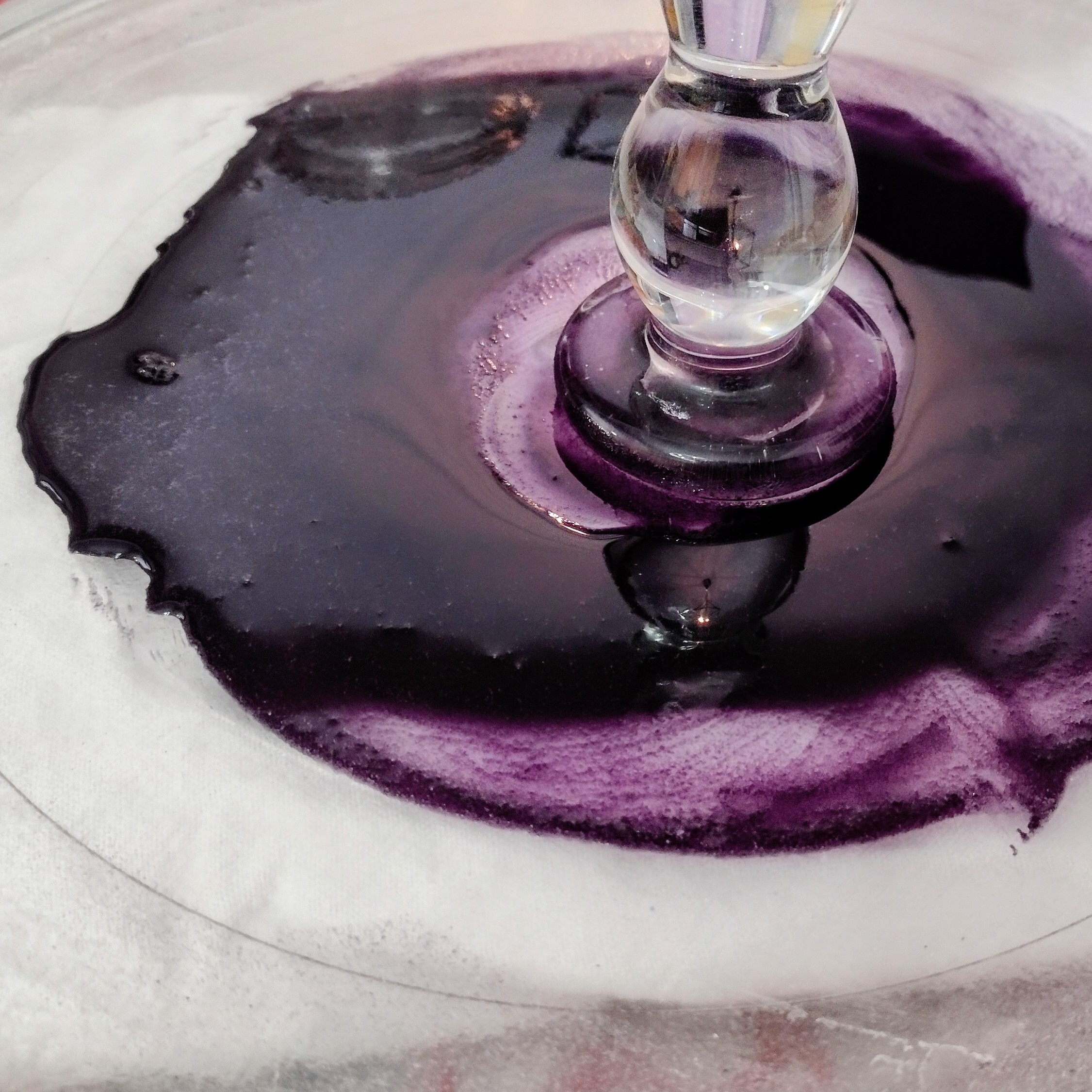 A glass platter with purple paint smeared in a pool on top. Sitting in the middle is a glass muller. 