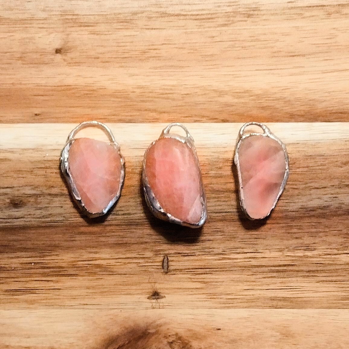 Three smooth rose quarts stones, each encircled with silver solder and loops at the top, on a wooden surface.