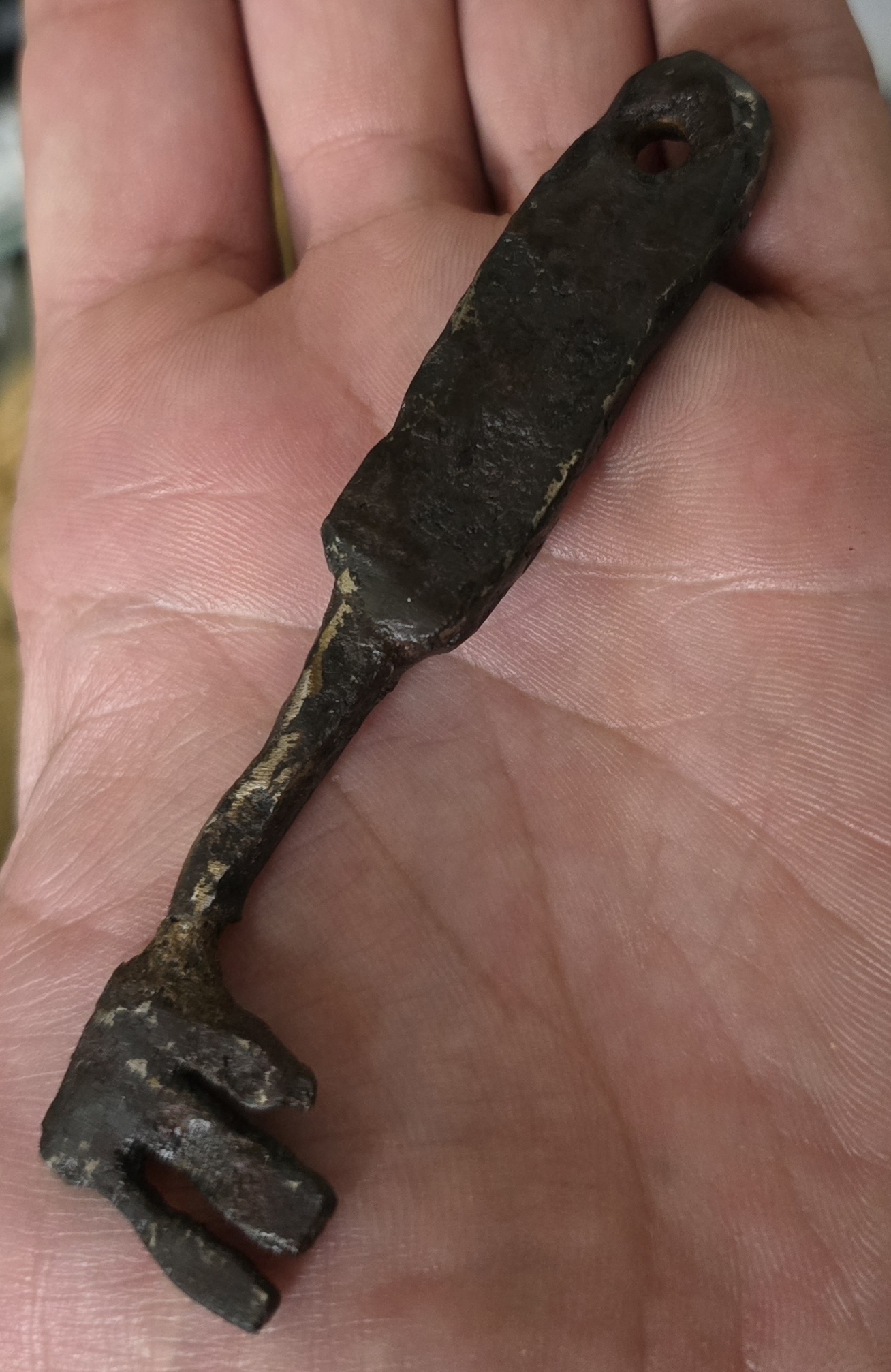 A rough-hewn, oxidized somewhat heavy bronze key with a rectangular shape and a hole in the upper end of the rectangle. The key tines are irregularly shaped but clearly distinguishable as serviceable.