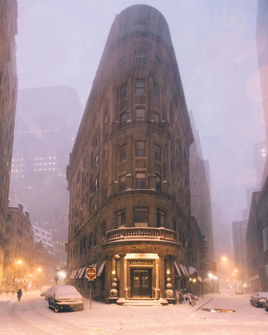 A tall wedge-shaped mid-rise building on the corner between two streets of New York. The air around is thick and grey with falling snow, and a warm homely light glows from lampposes in the two streets stretching away from the viewer. A lone figure strolls down the street to the left, and an office high-rise looms in the background. Other than that, the viewer is alone, and feels compelled to approach the Delmonico's building.