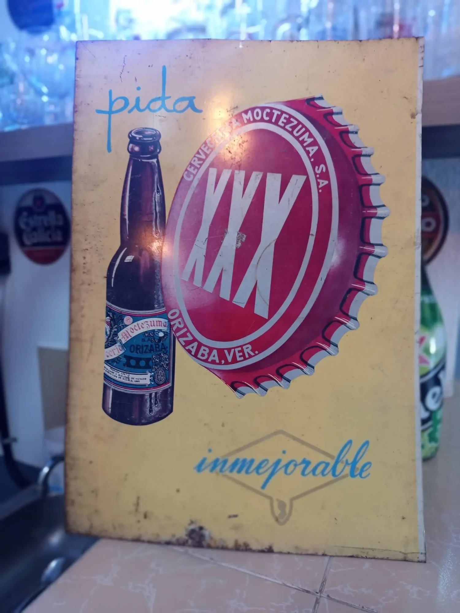 Metal sign advertising XXX beer: yellow bakground, left a bottle of XXX; centre, a large XXX bottle cap. Text: "pida" (order) & "inmejorable" (can't be bettered)