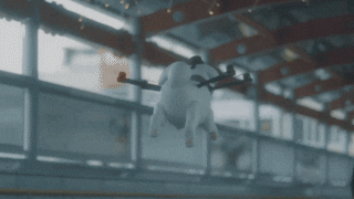 a GIF of a quadcopter drone shaped like a white dog with a tiny hat and doggy-paddling feet flying through the Yukimaru Road train station and Hakuba Shrine; the Japanese text overlays play like the flying dog is chatting with the townsfolk