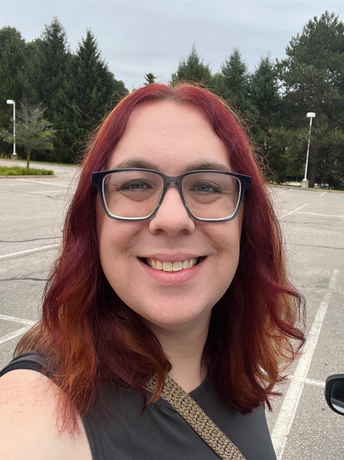 A white trans woman in her 30s with dyed red hair, wearing a dark gray tank top, is standing in a parking lot and smiling at the camera. She’s wearing glasses with a black rim and a purse with a tan knit strap. 