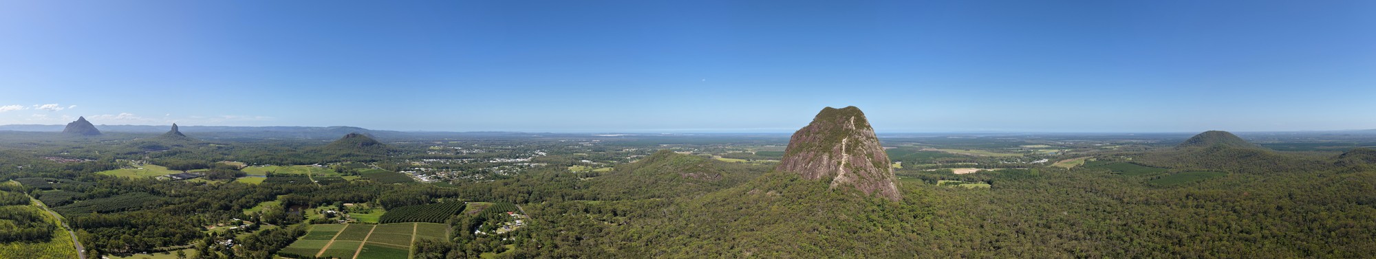 A panoramic drone photo showing the tree covered plains of the Glass House Mountains area, with the Glass House mountains themselves popping out here and there out of the landscape