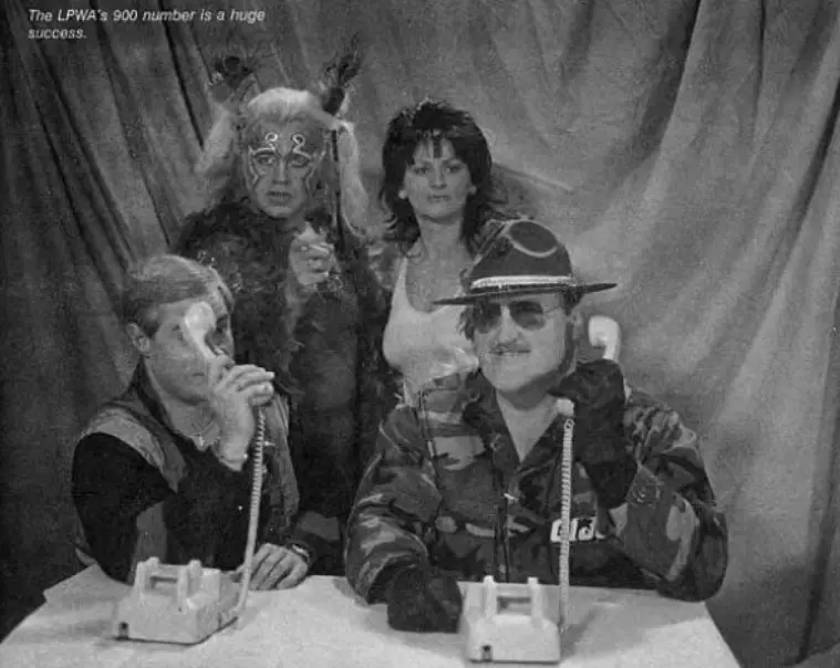 Sgt Slaughter and another dude sit at a table with large tabletop phones on them. Sarge stretches the receiver to his ear. The other dude is holding up his mid-gesture. Behind them Adrian Street and Miss Linda look like they're up to no good. Text in the top left reads, "The LPWA's 900 number is a huge success."