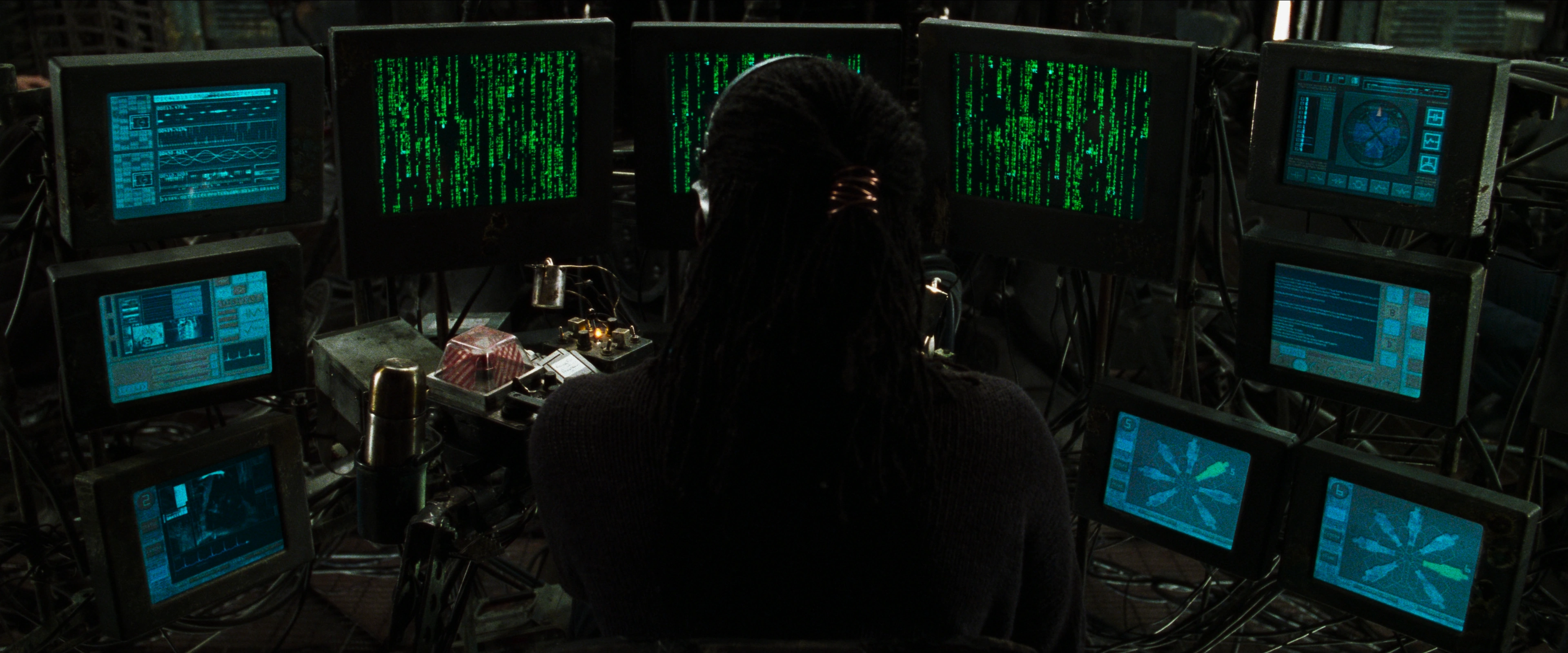 a screen capture from the 1999 film The Matrix showing the operator's desk with several monitors with black and green displays