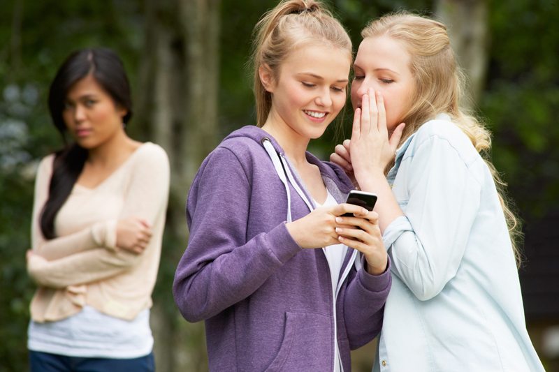 One girl whispers into the ear of another girl who is laughing and looking at her phone. In the background a third girl looks sad.