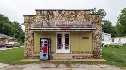 Photo of a grey day, an unabeled, likely abandoned store, about the size of a convenience store, with stone walls, old plywood where the sign once was, white doors with open spaces, and a pepsi machine. uncertain if the pepsi machine works.